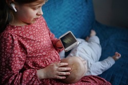 The older sister is watching the baby sister, and uses the phone with headphones. Close up older sister generation Z using phone, out of focus newborn sister.