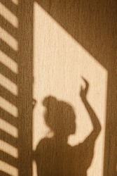 Silhouette of young woman stands with her hand up dancing in sunshine in the morning at home. Contrast window shadows on the wall. Aesthetic shade portrait. Lights and shades. Shade silhouette.