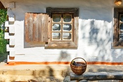 A wicker basket with an clay jug stands in the courtyard on a wooden bench near an old window with wooden shutters of a Ukrainian rural hut under a thatched roof in the sun.
