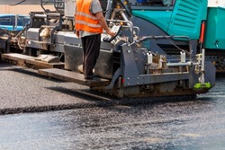 A road worker in an orange vest operates an asphalt paver and places hot fresh asphalt on a bitumen pavement while standing behind a control panel on a metal step on a summer day. Close-up, copy space