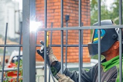 A worker welder in a protective helmet welds a metal grate to a fence against the backdrop of a brick house in blur.