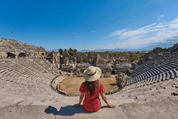 Tourist girl with hat and red dress posing, sitting and watching panorama of amphitheatre in ancient city of Side, Manavgat, Antalya, Turkey