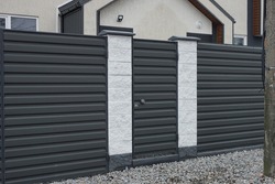 closed black metal door on a fence wall made of iron and white concrete pillars on a rural street