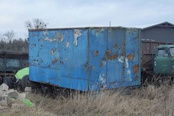 one big blue old iron trailer in  rust on the street overgrown with gray dry grass 