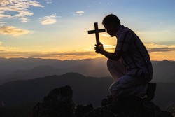 Silhouette Young man praying to the GOD while holding a crucifix symbol with bright sunbeam on top mountain