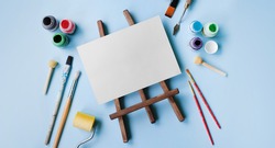 various tools for painting: brushes, spatula, roller, paints, blank on a wooden easel. flatley style on a blue background. copy space, mockup. art concept, learning to draw. baner style