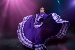 Latin woman dressed in a dress from Jalisco Mexico, purple skirmish with ribbons of bright colors, braids and in movement skirt and smile, black background