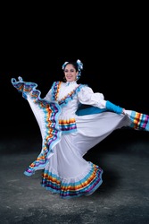 Latin woman dressed in a dress from Jalisco Mexico, white skirmish with ribbons of bright colors, braids and in movement skirt and smile, black background