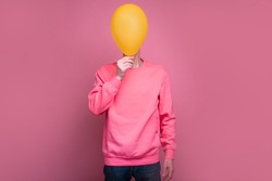Portrait full lenght of man in pink sweater cover face behind yellow balloon. Tall incognito male person stand alone. Isolated over pink background