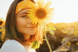 Lovely cheerful young woman posing alone on camera. Cover part of face with yellow sunflower blossom. Femlae model posing alone in middle of sunflower field. Morning or evening. Summertime