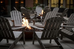 Low angle view of a fire pit surrounded by a circle of Adirondack chairs, with another campfire and hotel building in the dark background