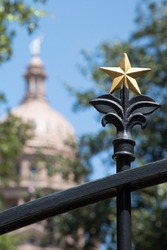 Close up on a decorative gold lone star on a wrought iron fence, with a spire dome building in the blurry background