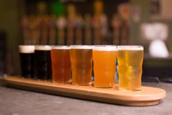 Low angle perspective close up of craft beer tasting flight at local brewery of small pint glasses in row on wooden tray with rainbow variety of golden yellow hoppy ales to dark malt stouts on bar