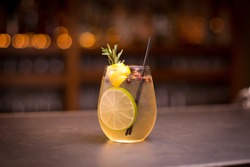 Low angle close up of ice cold modern gourmet craft cocktail of gin and tonic soda garnished by lemon slice and rosemary sprig sprinkled by juniper berries on bar with blurry restaurant bar background