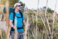 Portrait of a hiker, adult, male, Caucasian, with a backpack, mat, walking sticks, wearing a blue T-shirt, cap and gray pants, wearing a hearing aid and glasses.