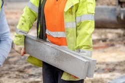 Close up of groundworker in orange and yellow hi-viz  carrying heavy concrete kerbs on construction site during new road construction