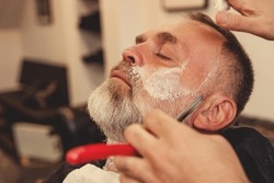 Shaving with a straight razor in a barbershop. A bearded old man being shaved in a barbershop. Classic shave by Stainless Steel Straight Edge Razor.