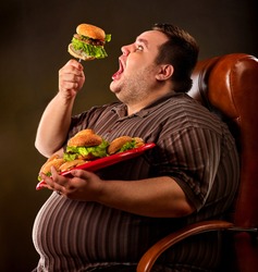 Diet failure of fat man eating fast food hamberger. Happy overweight person with wide-open mouth greedily eating huge hamburger on fork. Hate to diets. Business chairs for fat people concept.