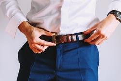 A man in blue pants and a white shirt buttoned a brown leather trouser belt. He has a watch on his hand