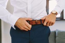 A man in blue pants and a white shirt buttoned a brown leather trouser belt. He has a watch on his hand.