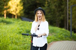 Trendy carefree woman with a surprised expression in white shirt and black hat ride on kick scooter outdoor in summer park street. Electric urban ecological transport concept.
