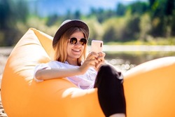 Portrait of charming smiling woman in sunglasses using cellphone and write messages to friends while lying on inflatable mattress on summer beach