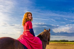 A pretty curly red haired attractive young Caucasian white woman looks at camera top down, riding a horse turns holding reins in sky clouds fields horizon. Horizontal day, summer outdoors copy-space