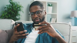 African young man in glasses shopping online with credit card using smart phone at home. Indoor.