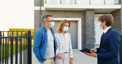 Caucasian young happy couple in medical masks buying house at outskirt and talking with male real-estate agent holding tablet device in hands. Outdoor. Coronavirus pandemic concept.
