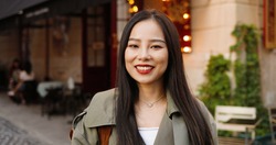 Portrait shot of pretty Asian happy young stylish woman looking and smiling to camera at nice street in town. Outdoors. Close up of beautiful cheerful female smile at cafe.