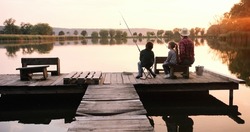 Little teen boy and girl with their grandpa sitting at the lake pier with rods and fishing equipment and talking early in the morning. Fishing and relationships concept 