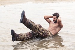 Handsome Muscled Soldier with no Shirt Doing Curl Ups Exercise at the Sea Water
