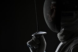 Profile of Young male fencer wearing white fencing Mask and costume holding the sword in front of his. Black Background