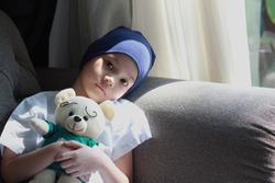 Patient kid lie down on couch or sofa in patient suit with her doll.Girl cover her head with  blue hat or headscarf.Kid look sad,tired and sick.Concept of   childhood cancer awareness.Selective focus.