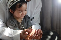 Religious Asian Muslim Man teaching his little son to pray to God with rosary at home.Sunset light shining through the window and touched boy body.Peaceful and Marvelous warm climate.Selective focus.