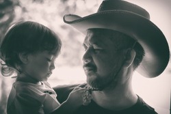 Monochrome retro film color tone of father with beard wearing cowboy hat and holding his kids in arms. Back lighting with grain.Vintage family portrait concept.