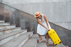 Beautiful tourist girl travels. Pulls a large yellow suitcase up the steps. Heavy load.