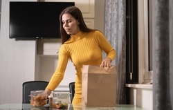 A beautiful Caucasian girl at home in the evening at home takes disposable lunch boxes with food from a paper bag and puts them on the table. Takeaway food concept. Copy space.