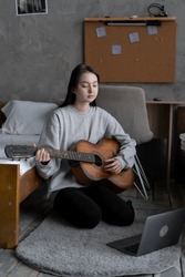 Asian teen girl playing guitar with laptop computer, learn to play with an online course in her room, sitting on the floor in casual clothes at home. Copy space