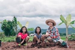 Mother and kids planting tree in garden, family Asian people working in farmland on hill and sky background, mom and children smile and holding seeding tree and dig soil for planting on dirt 