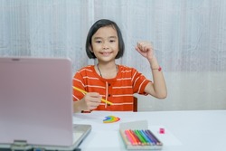 Kids learning and working online in room of house, happy Asian children smile and holding crayon or colored pencil to painting paper of homework, girl studen learning online with laptop computer