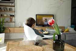 Little boy in feeding chair eating apples and bananas for breakfast in front of the cartoons on tv in a modern living room 