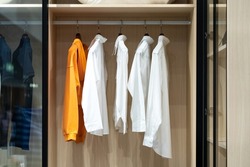 Shirts hang in a walking closet with glass door. One of those is in orange standout from others that are in white.     