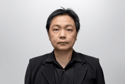 Asian man in his middle age, dressed in a stylish black suit and shirt, poses against a neutral white-gray backdrop.