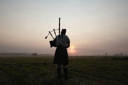 A musician plays the bagpipes in the fields at sunset.