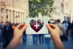 Health and wellbeing global issue as human hands holding a paper sheet with heart and cross icon over a crowded street background. Healthcare medical insurance, good life concept.
