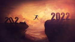 Determined man jump over a chasm obstacle to reach the new 2022 peak and let 2021 behind. Conceptual and surreal sunset scene, new year motivational background. Leader overcoming hurdles reach highs