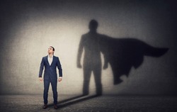 Determined businessman stands confident in a hero stance and casting a brave superhero shadow on the wall behind. Business leadership and motivation concept. Ambition and strength symbols