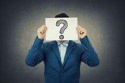 Businessman covering his face using a white paper with drawn question mark, like a mask, for hiding his identity. Isolated gray wall background.