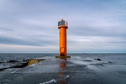 An orange lighthouse at the end of the Mangalsala or Eastern pier in Riga, Latvia where the river Daugava flows into the Baltic Sea. Winter storm, cold weather, ice and snow.